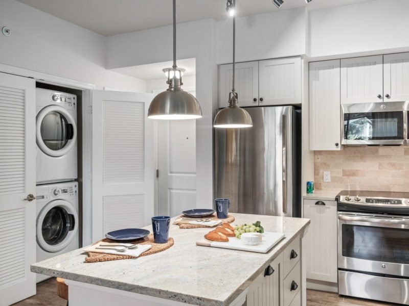 This image shows an expansive view of the Premium Apartment Feature, especially the kitchen island showcasing a neat granite-inspired countertop, accessible kitchen pieces of equipment, and an aisle directly through washer and dryer area.